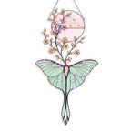 A hanging stained-glass style ornament shaped like a large pale green moth with a pink circular sun and small pink flowers above it, outlined in dark metal and shown on a white background.