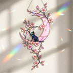 A hanging stained-glass style ornament featuring a black cat sitting on a pink crescent moon with pink cherry blossom branches and iridescent floral accents, shown against a softly lit neutral background with rainbow light streaks.