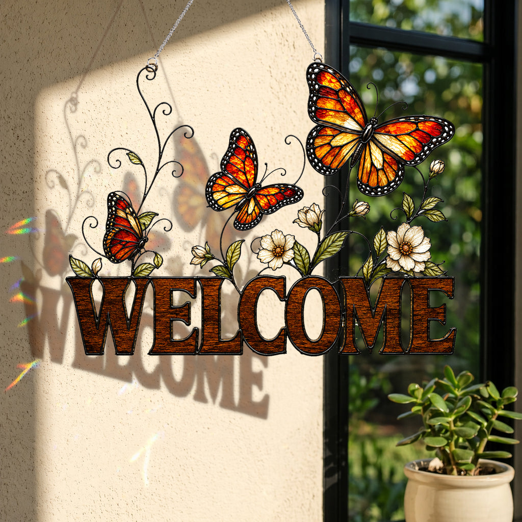 A decorative hanging 'WELCOME' sign with three orange and yellow butterflies, white flowers, and black scroll vines, displayed in front of a window with sunlight casting shadows on a beige wall.