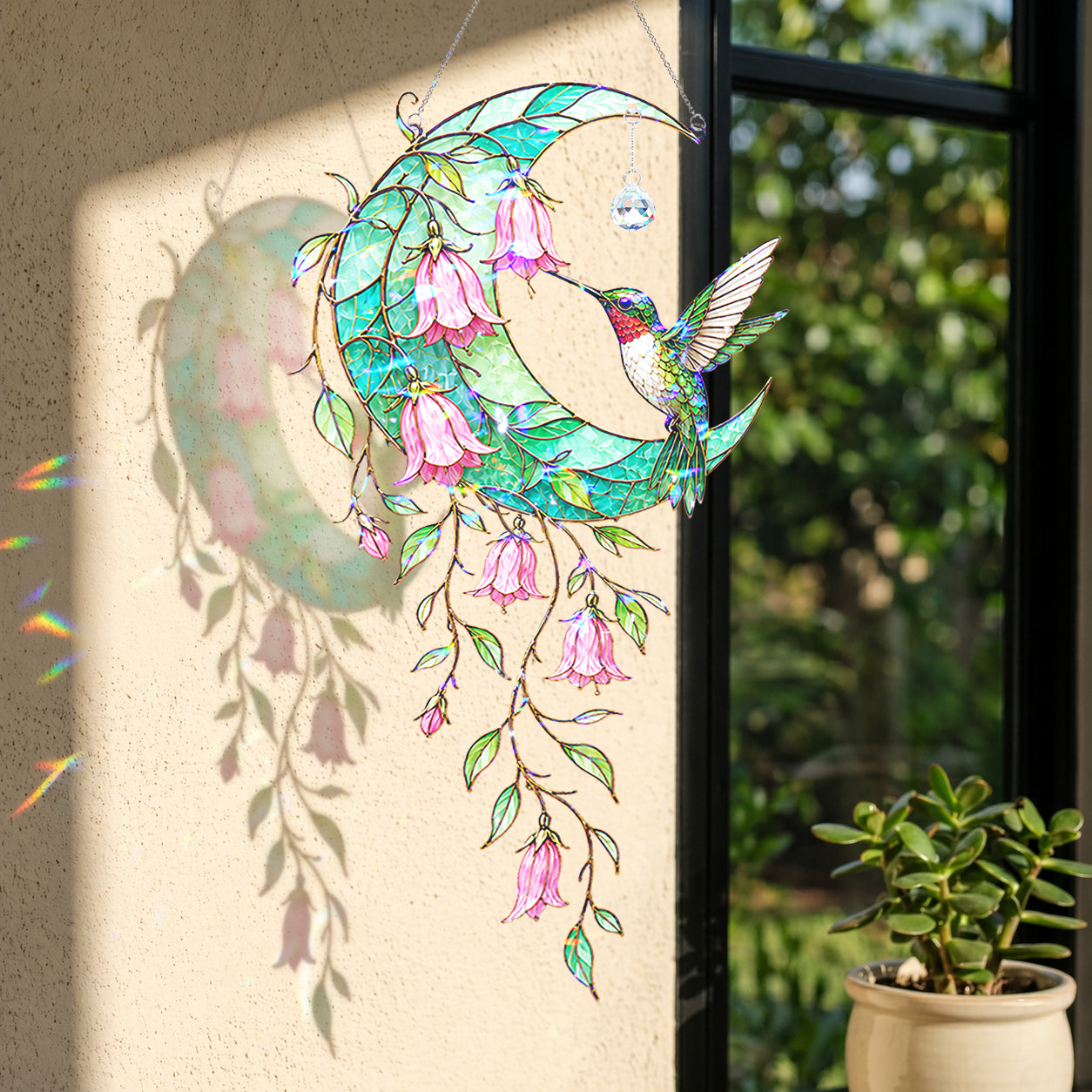 A stained-glass style hanging ornament featuring a crescent moon, hummingbirds, and pink bell-shaped flowers, displayed in front of a window with sunlight casting a shadow on the wall.