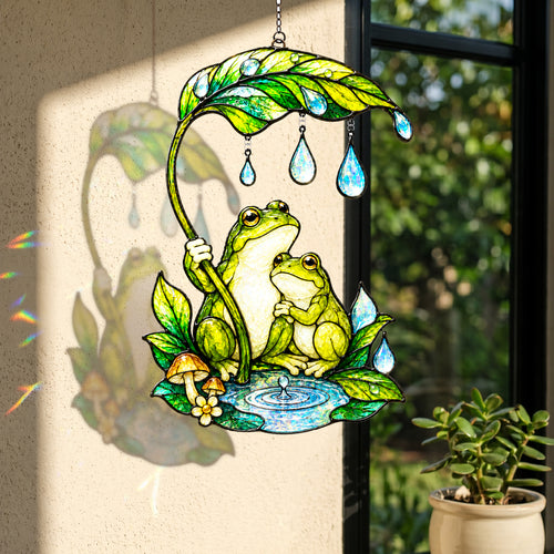 A stained-glass style hanging ornament featuring two green frogs under a large leaf with teardrop-shaped blue drops, displayed indoors near a window and casting a shadow on the wall.