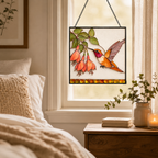 Hummingbird fuchsia stained glass suncatcher hanging in a bedroom window beside soft bedding, a wooden nightstand, books, and a candle.