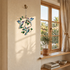 Hummingbird Leaf holographic acrylic suncatcher hanging on a warm sunlit wall beside a wooden window and potted plant.