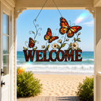 A decorative hanging sign with the word “WELCOME” in dark brown letters, decorated with three orange-and-black butterflies, white flowers, and green leaves, displayed on a porch with a beach and ocean background visible through the open doorway.