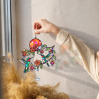 Hand holding a colorful stained glass sun catcher with hummingbirds and flowers against a window.