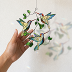 Close-up of a hand holding a Hummingbird Leaf holographic acrylic suncatcher, showing three hummingbirds, green leaves, and rainbow light reflections.