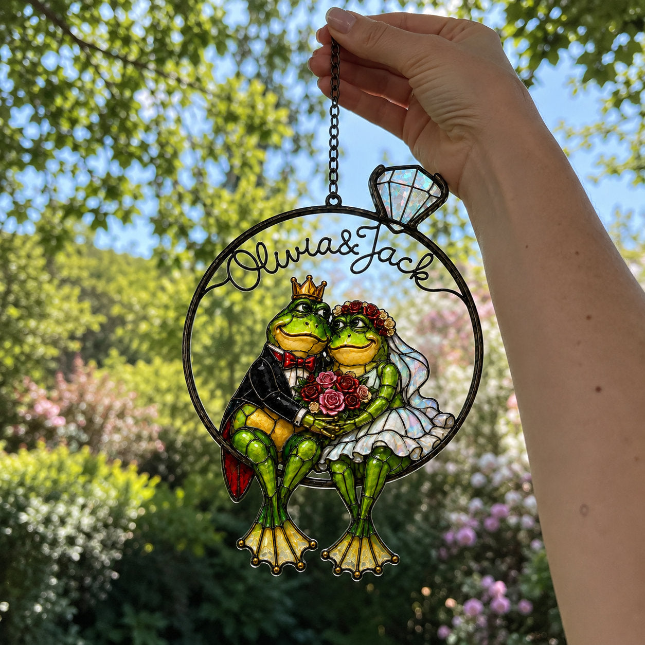Hand holding the personalized frog wedding couple ring suncatcher by its chain outdoors