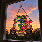 Frog couple scooter suncatcher in a window against a pink sunset sky