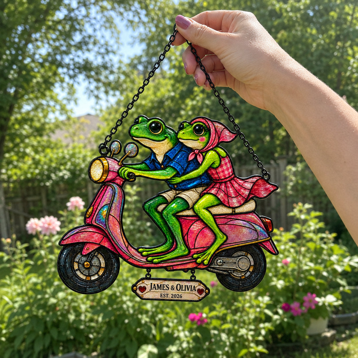Hand holding the personalized frog couple scooter suncatcher by its chain outdoors