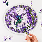 Hand placing a puzzle piece into the Amethyst Bellflower Hummingbird acrylic puzzle suncatcher with loose pieces nearby.
