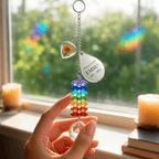 Hand holding personalized birth flower crystal suncatcher near a sunny window with rainbow reflections, marigold heart charm, custom Emma teardrop charm, and crystal ball
