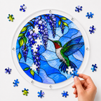 Hand placing a puzzle piece into a blue wisteria hummingbird acrylic puzzle suncatcher with loose blue and purple pieces around it