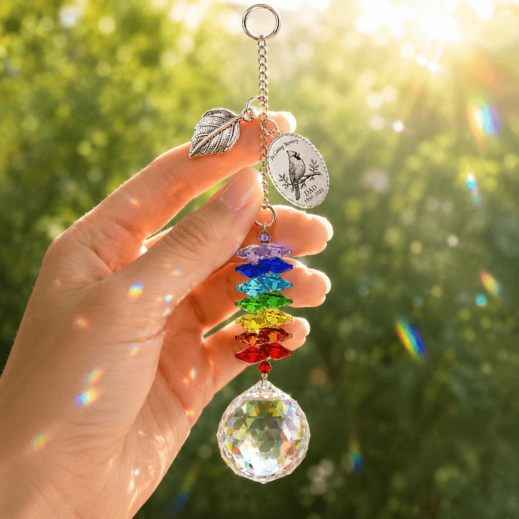 Hand holding personalized cardinal memorial crystal suncatcher near a sunny window with leaf charm, custom oval charm, rainbow crystals, and crystal ball