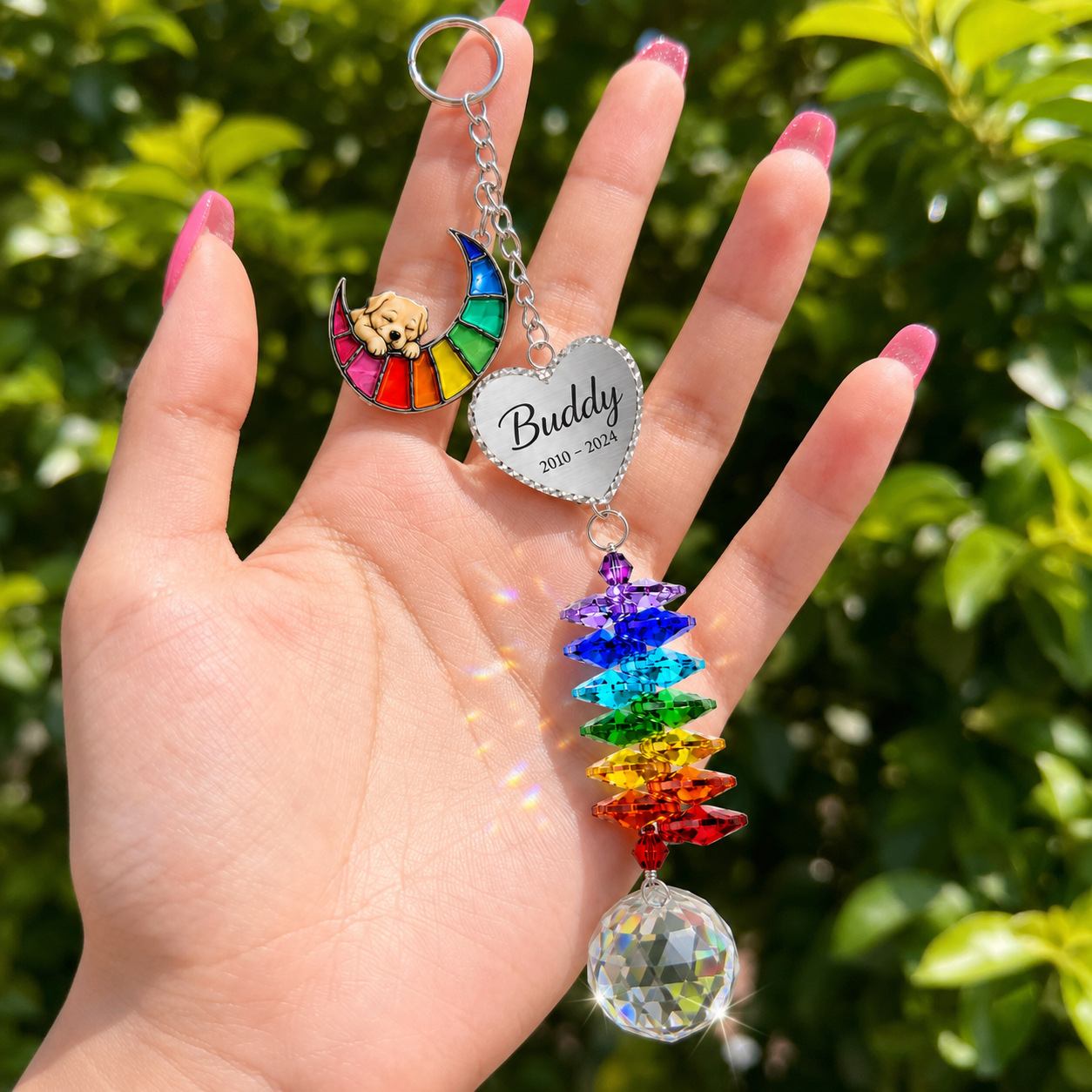 Hand holding personalized dog memorial Rainbow Bridge crystal suncatcher outdoors with green garden background and sunlight