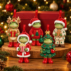 Five Dressed for Christmas frog ornaments standing on a wooden board in front of a red gift box and a lit Christmas tree