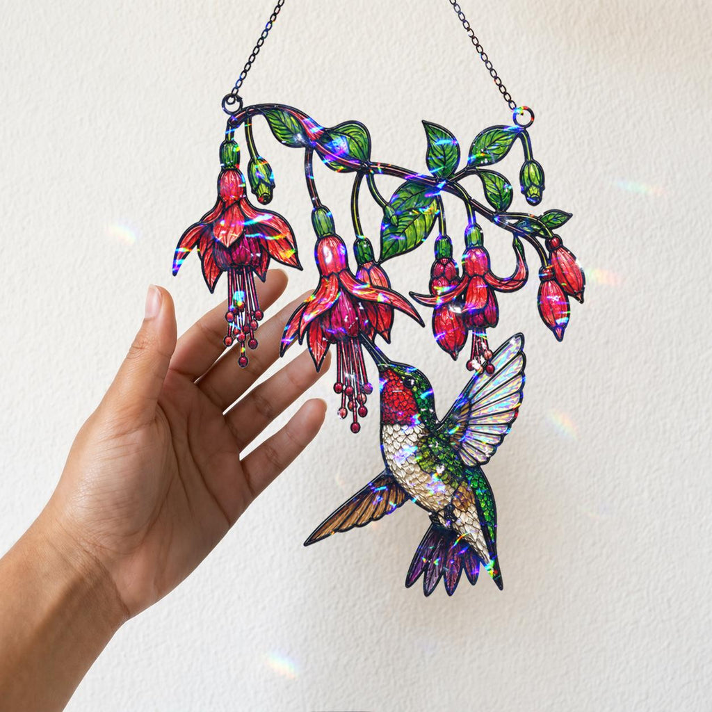 A hand holding the hanging chain of the Fuchsia Hummingbird holographic acrylic suncatcher near a warm wall with soft sunlight and rainbow reflections.