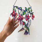 A hand holding the hanging chain of the Fuchsia Hummingbird holographic acrylic suncatcher near a warm wall with soft sunlight and rainbow reflections.