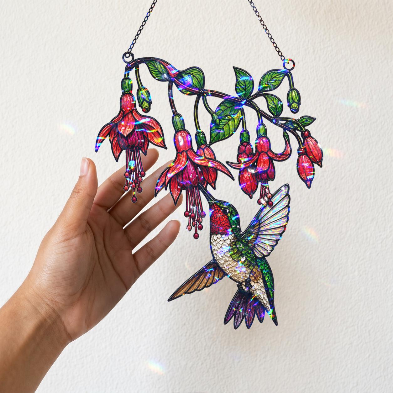 A hand holding the hanging chain of the Fuchsia Hummingbird holographic acrylic suncatcher near a warm wall with soft sunlight and rainbow reflections.