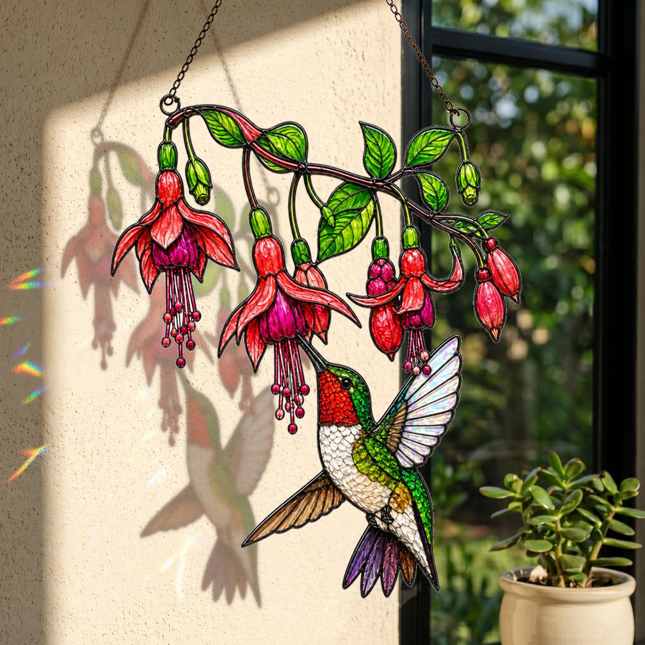 Colorful hummingbird and fuchsia flower window suncatcher displayed beside a sunlit indoor plant.