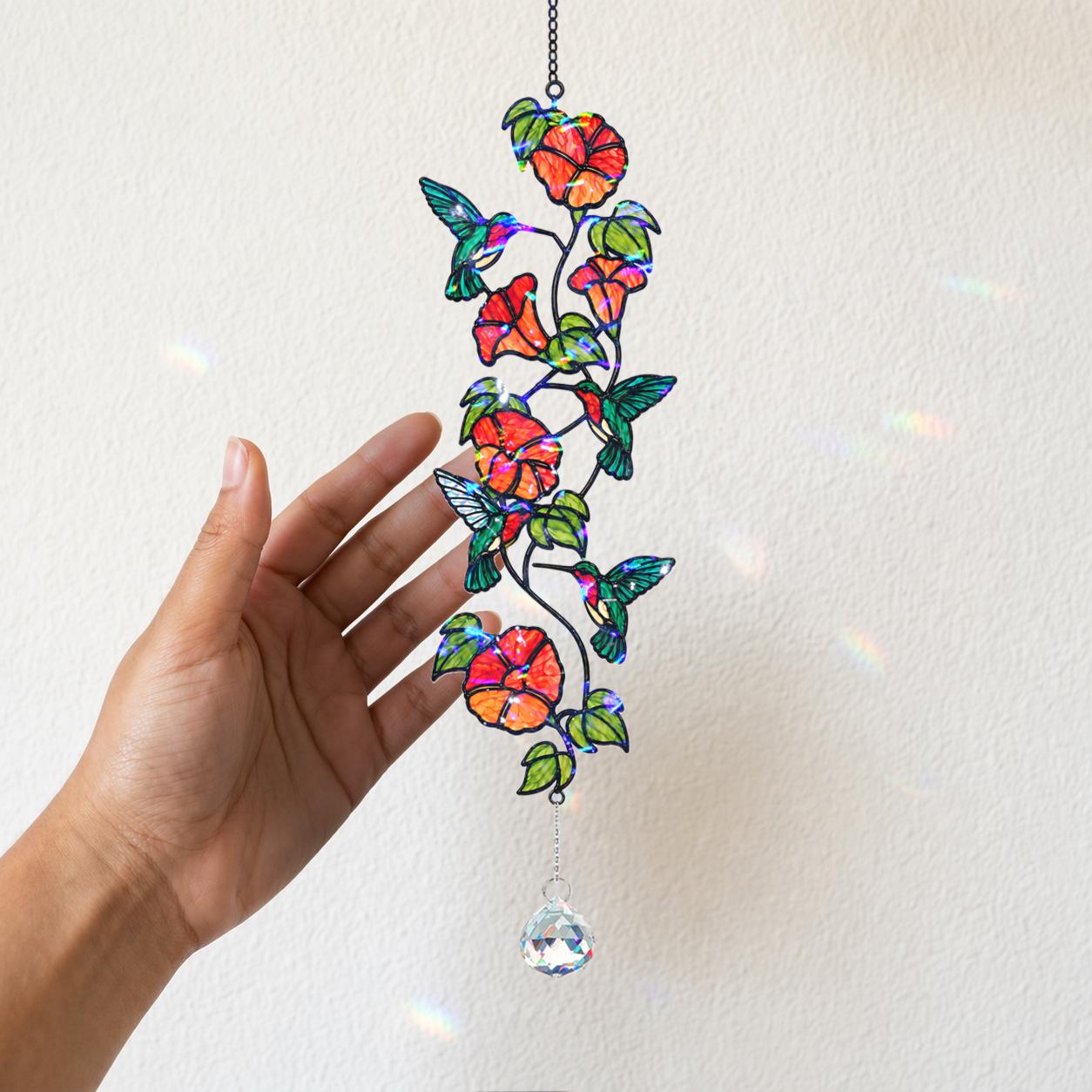 Hand holding a vertical hummingbird and orange flower holographic acrylic suncatcher against a white wall.
