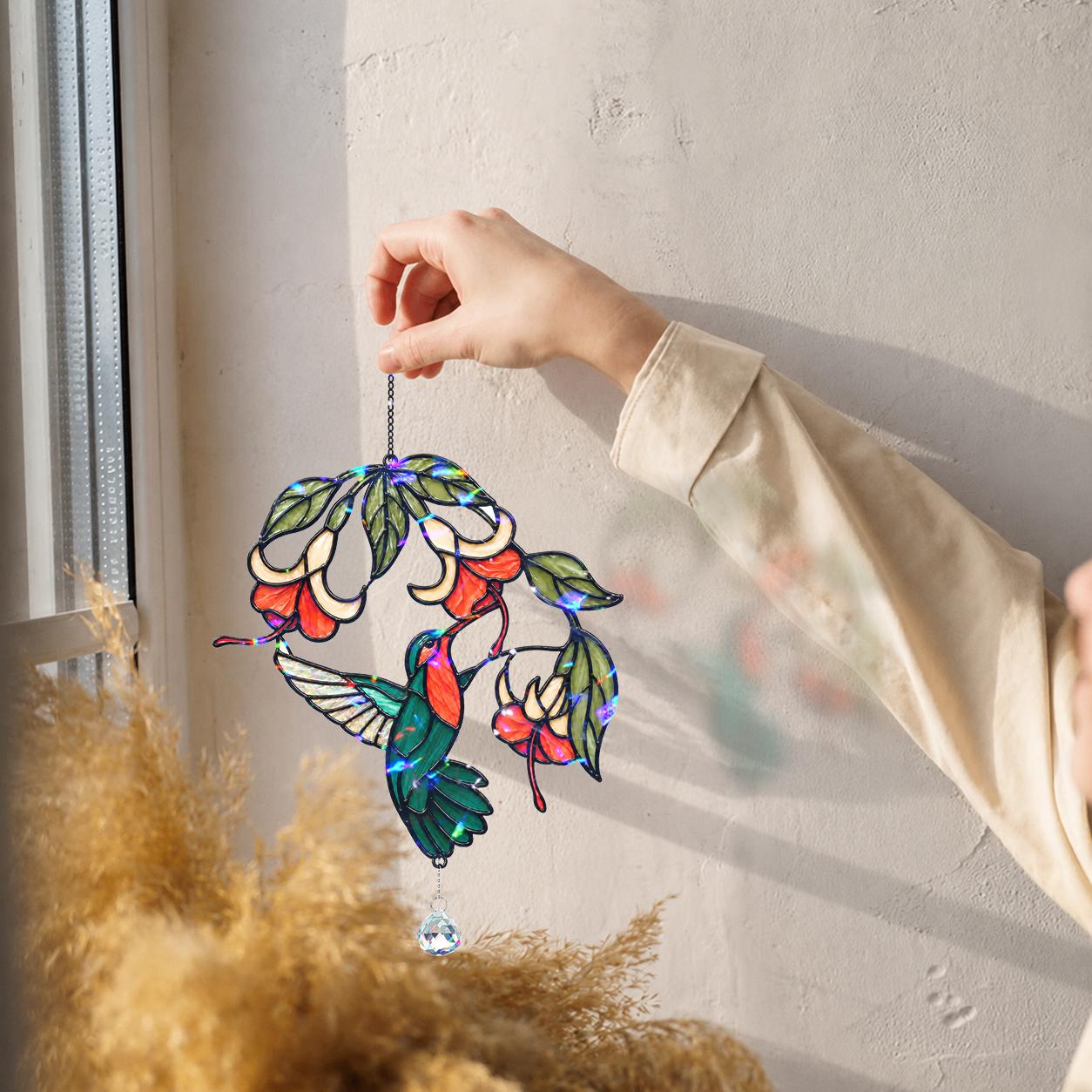 Hand holding a colorful hummingbird fuchsia flower holographic acrylic suncatcher near a sunny wall with soft shadows.
