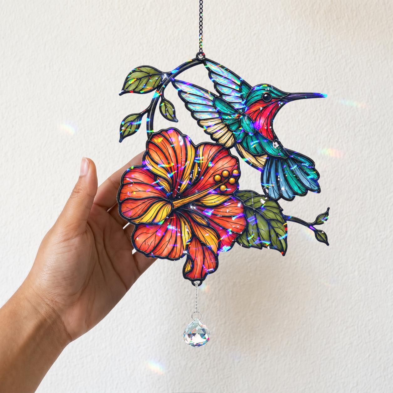 Hand holding a colorful hummingbird hibiscus holographic acrylic suncatcher near a sunny wall with soft shadows.
