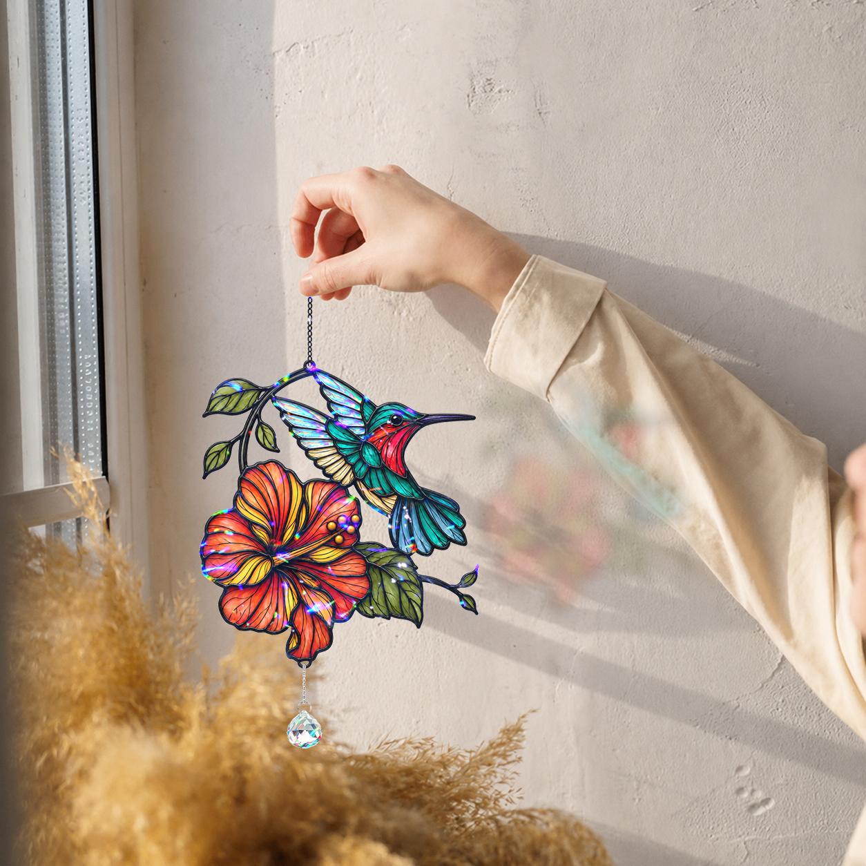 Hand holding hummingbird hibiscus holographic acrylic suncatcher against a white wall for size reference.
