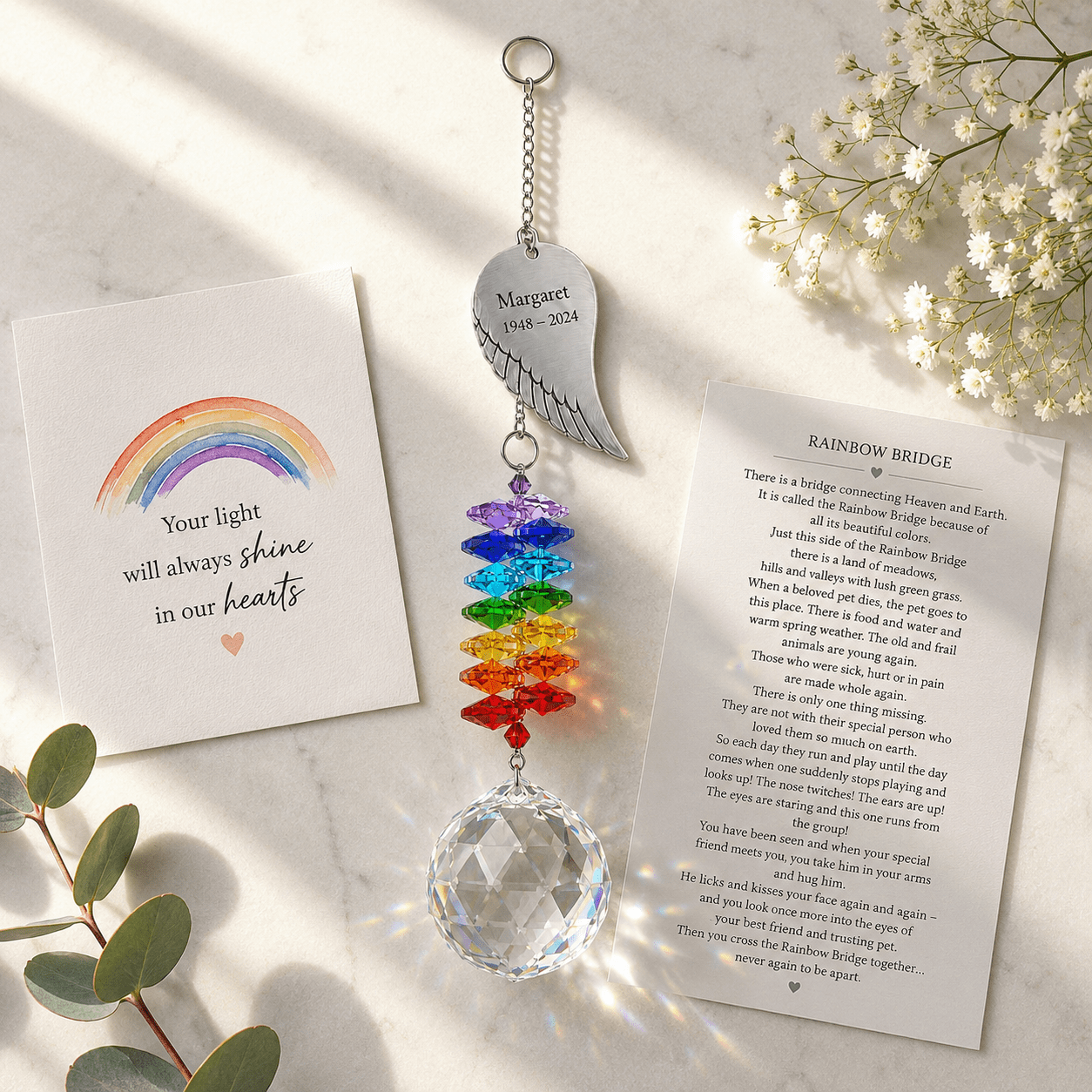 Personalized memorial angel wing crystal suncatcher displayed on marble surface with Rainbow Bridge remembrance card and soft sunlight