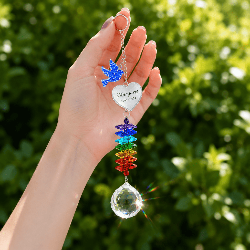 Hand holding personalized memorial dove crystal suncatcher outdoors with blue dove charm, custom heart charm, rainbow crystals, and sparkling crystal ball