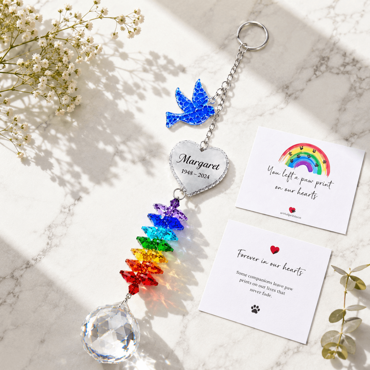 Personalized memorial dove crystal suncatcher displayed on marble surface with sympathy cards, rainbow crystals, and white flowers