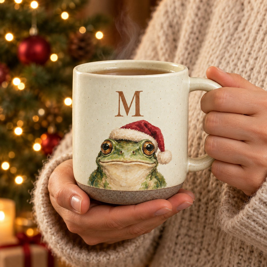 Hands in a cream sweater holding the initial M frog pottery mug in front of a lit Christmas tree