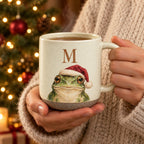 Hands in a cream sweater holding the initial M frog pottery mug in front of a lit Christmas tree