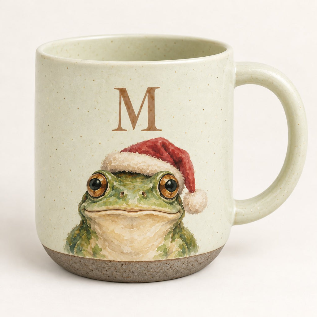 Front view of the frog pottery mug with the initial M: speckled cream stoneware, raw clay base, watercolor frog in a Santa hat