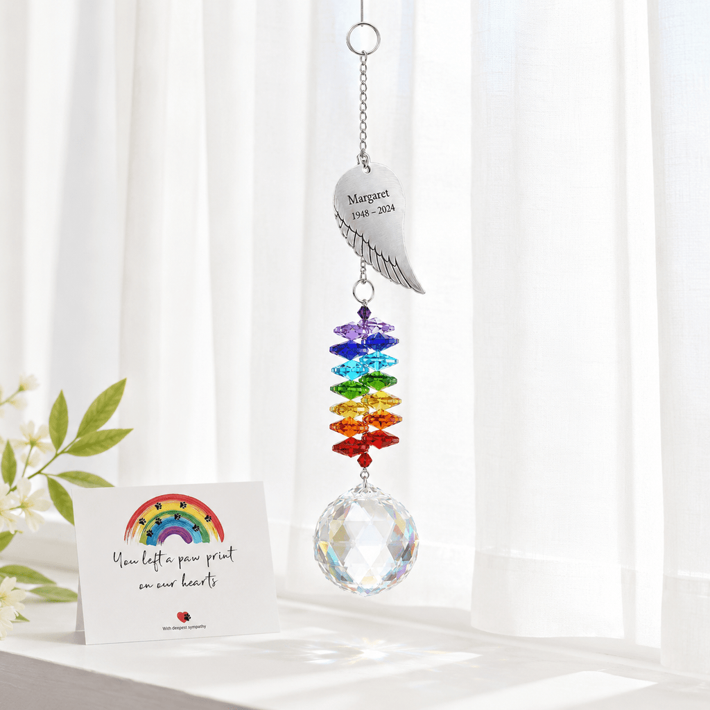 Personalized angel wing memorial crystal suncatcher hanging in front of sheer curtains with remembrance card and white flowers