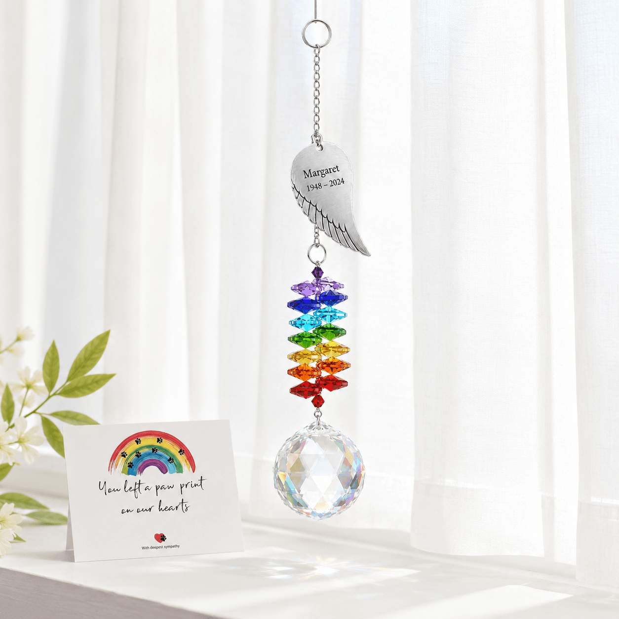Personalized angel wing memorial crystal suncatcher hanging in front of sheer curtains with remembrance card and white flowers