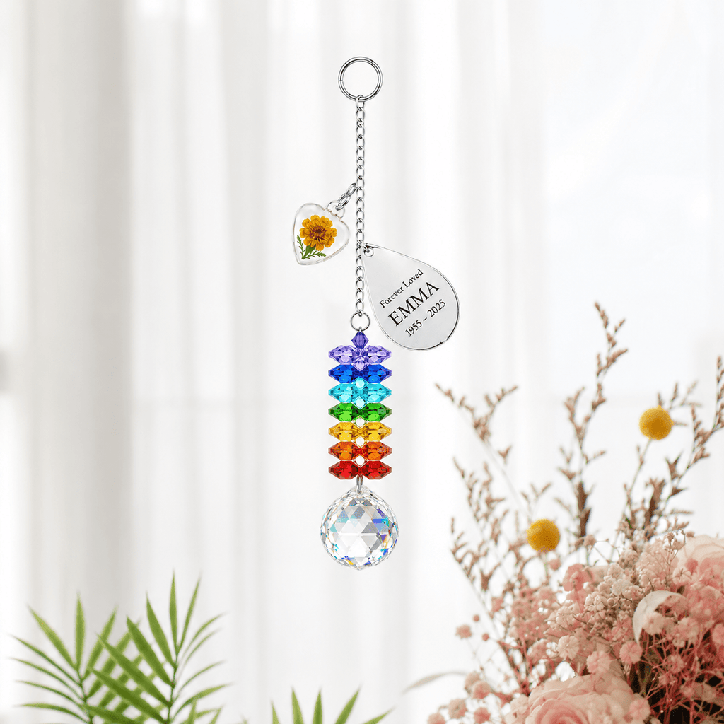 Personalized birth flower crystal suncatcher hanging in front of soft white curtains with marigold heart charm, custom Emma teardrop charm, rainbow crystals, and clear crystal ball