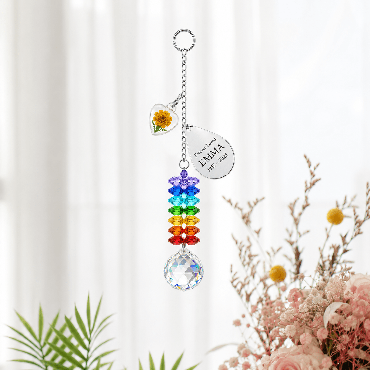 Personalized birth flower crystal suncatcher hanging in front of soft white curtains with marigold heart charm, custom Emma teardrop charm, rainbow crystals, and clear crystal ball