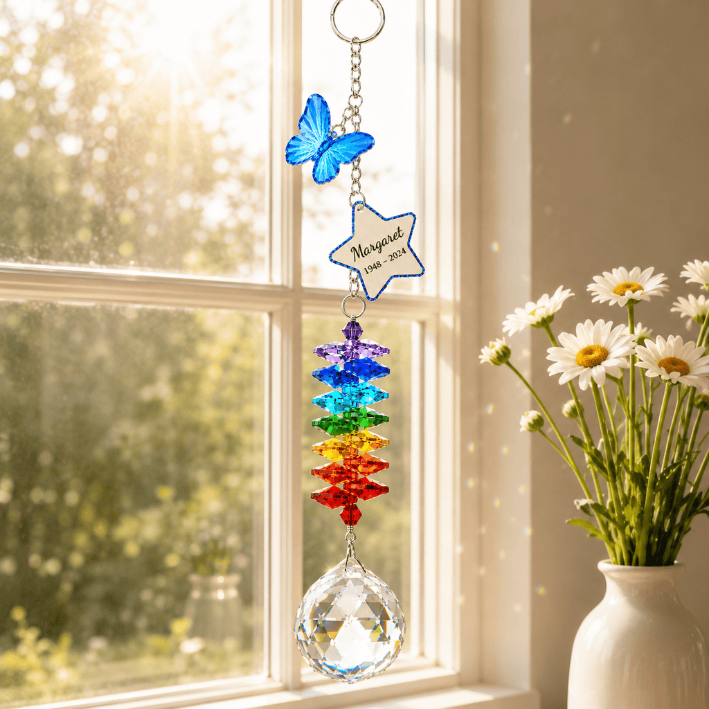 Personalized butterfly memorial crystal suncatcher hanging in front of sheer curtains with Rainbow Bridge sympathy card and white flowers