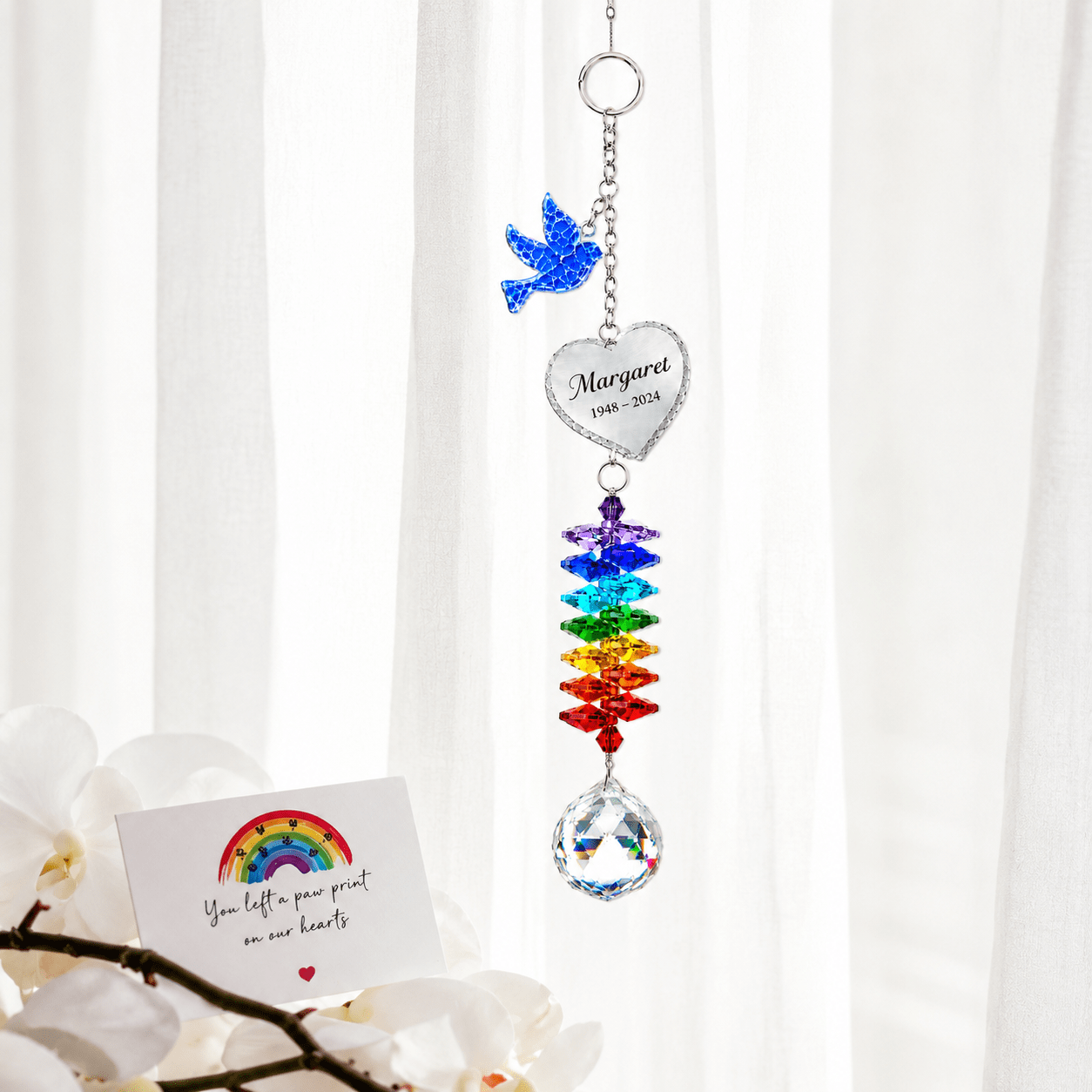 Personalized memorial dove crystal suncatcher hanging in front of sheer curtains with sympathy card and white orchid flowers