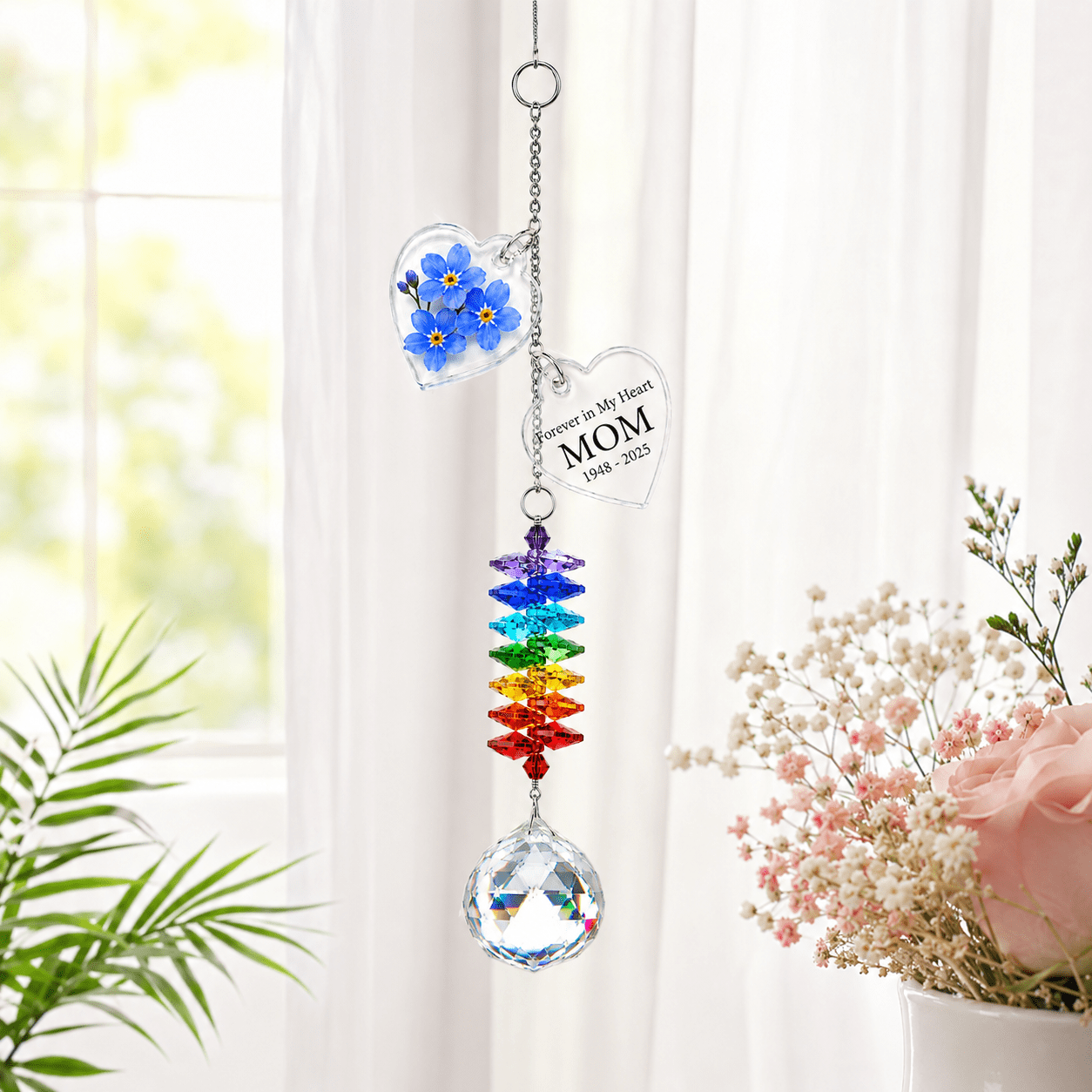 Personalized memorial flower crystal suncatcher hanging by a bright window with blue forget me not charm, custom star charm, rainbow crystals, and soft floral decor