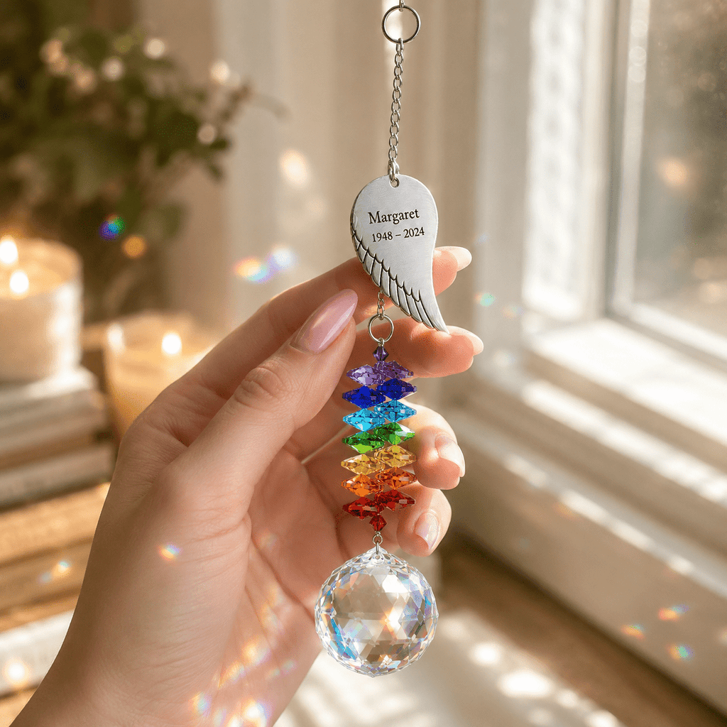 Hand holding personalized memorial angel wing crystal suncatcher near a sunny window with rainbow crystal reflections