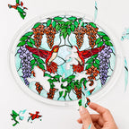 Hand placing a puzzle piece into the Scarlet Duet Hummingbird Blossom acrylic puzzle suncatcher with loose pieces nearby.