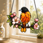 Stained glass style suncatcher with a Baltimore oriole on a branch of pink apple blossoms, hanging in a sunlit window with rainbow light