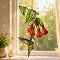 Stained glass style suncatcher with a hummingbird under hanging red trumpet flowers, hanging in a sunlit window with rainbow light