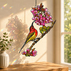 Stained glass style suncatcher with a scarlet sunbird under a cluster of pink blossoms, hanging in a sunlit window with rainbow light