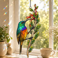 Stained glass style suncatcher with a rainbow sunbird feeding at red tubular flowers, hanging in a sunlit window with rainbow light