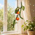 Stained glass style suncatcher with two green tree frogs climbing a vine with orange berries, hanging in a sunlit window with rainbow light