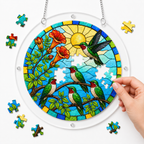 Hand placing a puzzle piece into a sunlit hummingbird family trumpet vine acrylic puzzle suncatcher with colorful loose pieces around it