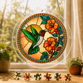 Round Tangerine Bloom Hummingbird acrylic puzzle suncatcher hanging in a sunny window with colorful loose puzzle pieces below.