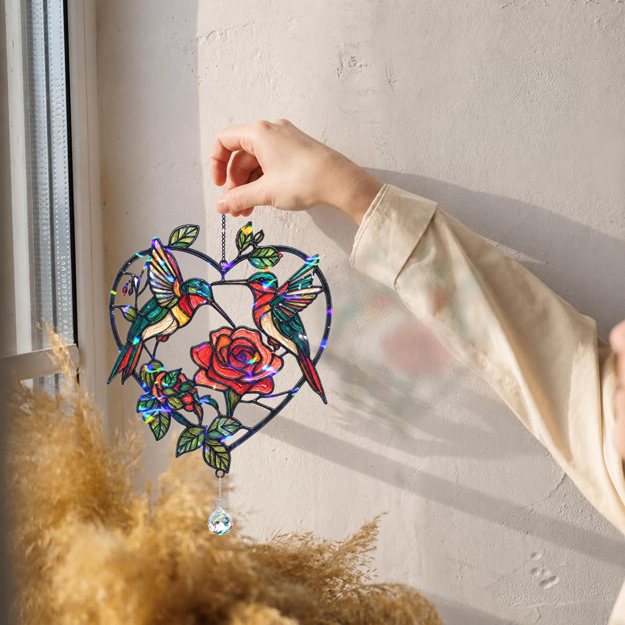 Hand holding a two hummingbirds rose heart holographic acrylic suncatcher near a sunny wall with soft shadows.
